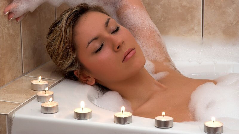 Junge Frau liegt im Schaumbad in Badewanne mit Teelichtern auf dem Rand, geschlossene Augen, terrakotta-farben