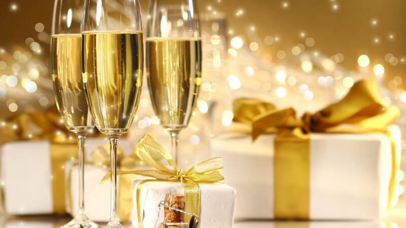 Drei Gläser Champagner, daneben weiß verpackte Geschenke mit goldener Schleife, dahinter goldenes Feuerwerk