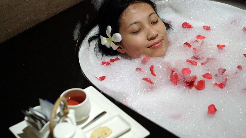 Junge Frau entspannt im Schaumbad mit Rosenblättern, neben ihr Tablett mit Tee