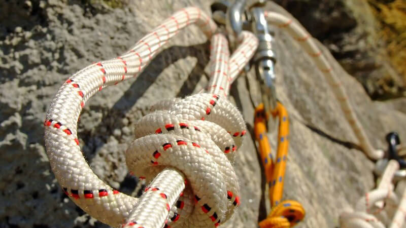 Nahaufnahme Bergsteigersicherung an Berg befestigt, daran weißes und orangenes Seil