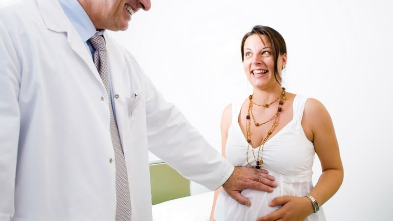 Arzt mit weißem Kittel hält schwangerer Frau in weißem Kleid Hand auf Bauch