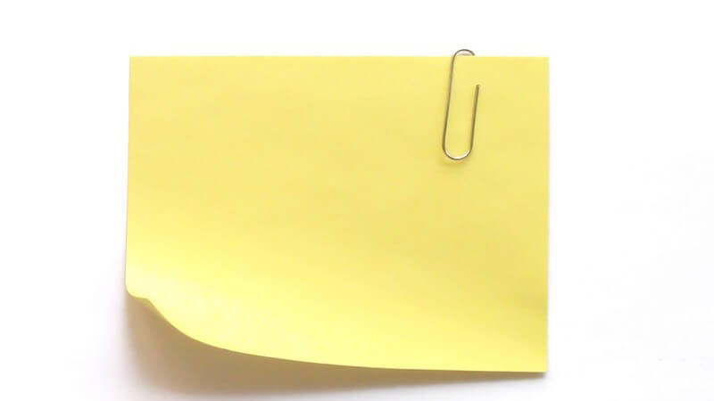 Gelber Post-It - Notizzettel mit Büroklammer aus Eisen mitte rechts vor weißem Hintergrund