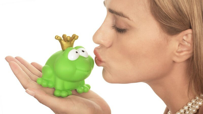 Frau mit Perlenkette um Hals küsst mit geschlossenen Augen grünen Plastikfrosch mit goldener Krone