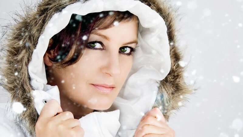 Frau mit weißer Kapuze mit Fell zieht Kragen hoch, steht im Schnee