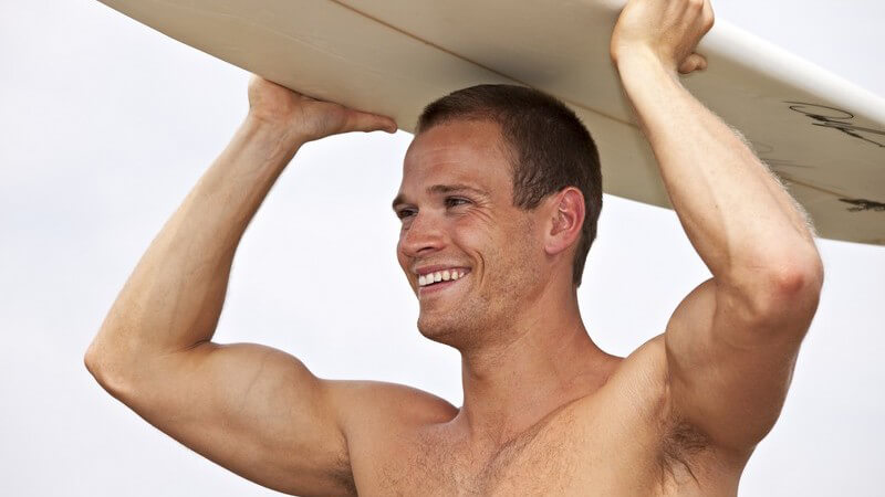 Junger Mann, oberkörperfrei, trägt mit beiden Händen Surfbrett auf dem Kopf gelehnt und lächelt